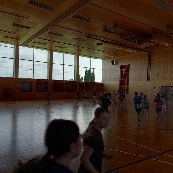 Ein Basketball-Hallenspielfeld mit mehreren spielenden Personen. Der Platz hat Holzböden und Glasfenster. Rechts steht ein Basketballkorb und links steht eine Person.