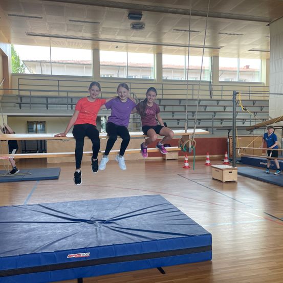 Drei junge Mädchen springen auf einer horizontalen Stange in einer Turnhalle. Eine trägt ein lila Shirt, eine andere orange und die dritte schwarz. Dahinter befindet sich eine Turnhalle mit einer blauen Matte, einem Holzkasten und orangefarbenen Verkehrskegeln.