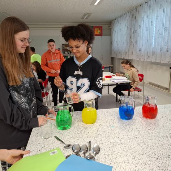 Zwei Mädchen stehen vor einer Arbeitsplatte mit bunten Flüssigkeiten in Gläsern und Krügen. Eine weitere Mädchen sitzt im Hintergrund an einem Tisch.