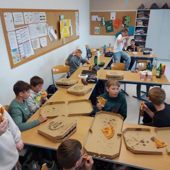 Eine Gruppe von Jungen isst Pizza in einem Klassenzimmer. Ein Tisch mit Pizzakartons und Bechern steht vor ihnen. Der Junge in der Mitte hält ein Pizzastück.