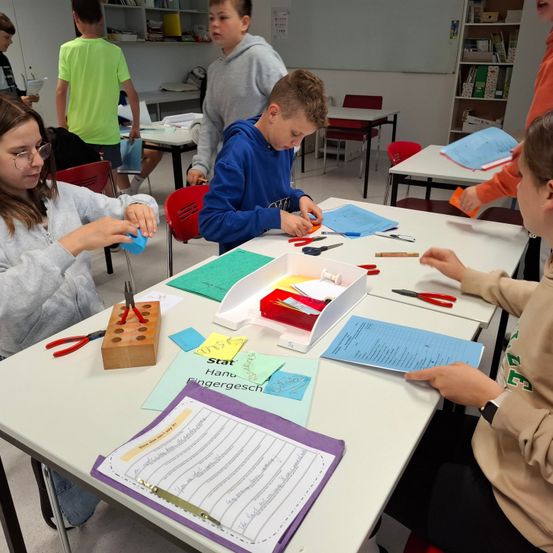 Schüler in einem Klassenzimmer arbeiten an einem Projekt. Ein Schüler benutzt eine Schere, um ein Stück Papier zu schneiden, und ein anderer hält ein blaues Objekt. Der Tisch hat eine Schachtel mit verschiedenen Werkzeugen und Papieren.