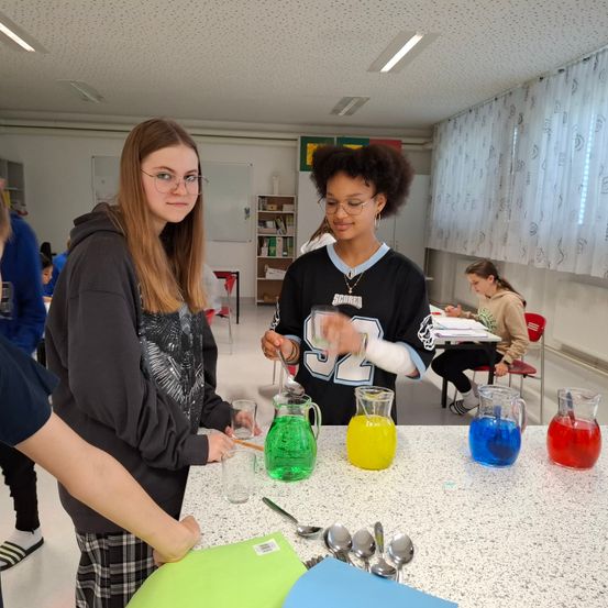 Zwei Frauen stehen vor einem Tisch mit bunten Flüssigkeiten in Krügen und Gläsern. Im Hintergrund sind andere Personen.