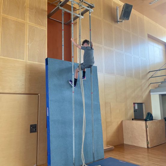 Ein junger Mann in einem grauen Shirt und Shorts klettert an einer blauen Wand mit einem Seil. Der Raum hat Holzböden, eine Holztür und eine Wand mit einem Lautsprecher.