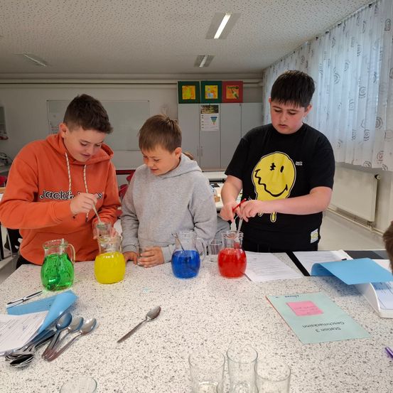 Drei Jungen führen ein Chemieexperiment mit farbigen Flüssigkeiten in Bechern durch. Sie schauen sich gegenseitig und den Inhalt der Becher an.