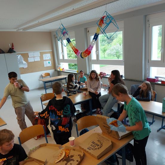 Eine Gruppe von Kindern isst Pizza in einem Klassenzimmer. Einige sitzen auf Stühlen, andere stehen.
