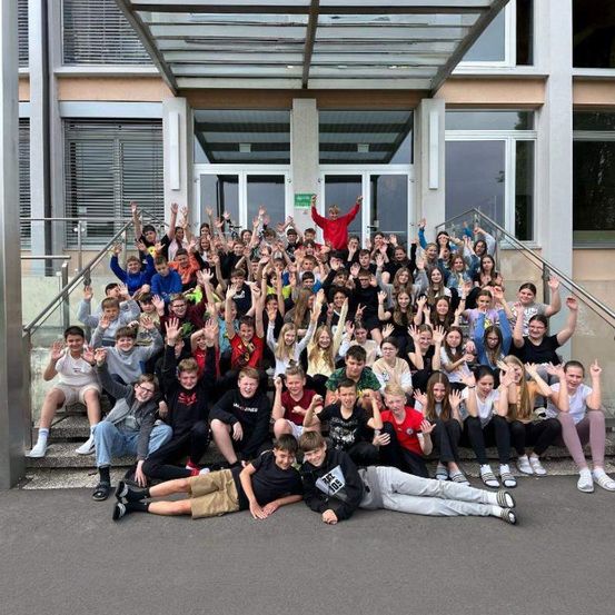 Gruppenfoto von Schülern einer Schule, die auf Stufen vor dem Schulgebäude stehen und sitzen und mit den Händen winken.