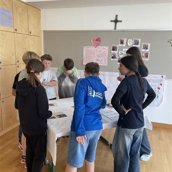 Eine Gruppe von Kindern versammelt sich um einen Tisch im Klassenzimmer, einige trinken aus Tassen. Eine Person steht am Tisch mit einem Buch, andere schauen an eine Tafel. Der Raum hat einen Holzboden und eine Pinnwand mit Bildern und Text.