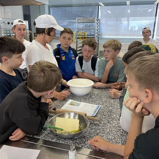 Eine Gruppe junger Jungen versammelt sich um einen Tisch in einer Küche und hört einer Frau mit einer weißen Kappe zu. Ein Topf, ein Nudelholz und ein Stück Papier liegen auf dem Tisch.