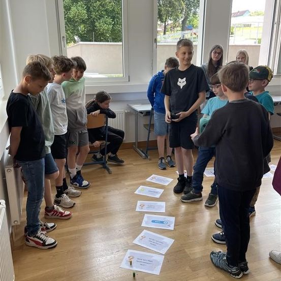 Eine Gruppe von Kindern steht in einem Kreis mit einigen Papieren auf dem Boden. Ein Junge hält eine Fernbedienung. Einige Kinder sitzen auf Stühlen. Ein Tisch mit einem Heizkörper steht neben ihnen. Bäume und ein Gebäude sind draußen.
