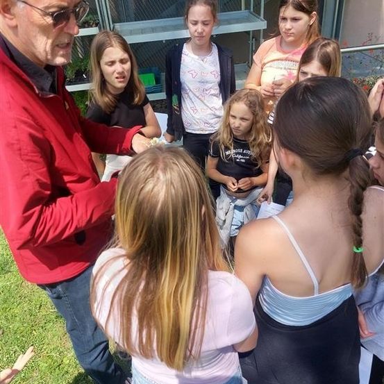 Ein älterer Mann mit einer Brille steht in einem Garten mit einer Gruppe junger Mädchen, sie scheinen in ein Gespräch vertieft zu sein. Einige der Mädchen schauen zu ihm.