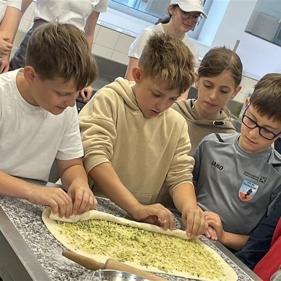 Mehrere Jungen sind in einer Küche und arbeiten an einem Pizzateig. Ein Junge benutzt einen Nudelholz. Ein anderer Junge schaut sich den Pizzateig an. Sie tragen alle Freizeitkleidung.