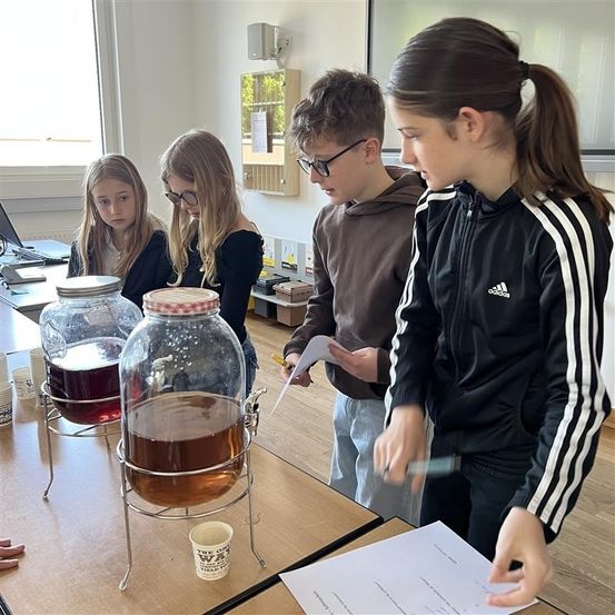 Vier Schüler stehen um einen Tisch herum, zwei Mädchen und zwei Jungen. Sie scheinen an einer Klassenaktivität teilzunehmen, mit zwei großen Teekannen in der Mitte. Ein Mädchen hält ein Papier und einen Stift, während ein anderes Mädchen in die Kamera schaut. Ein Junge schreibt auf ein Papier, und ein anderer Junge schaut darauf.