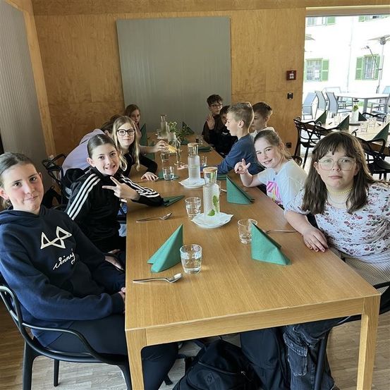 Eine Gruppe von Menschen, möglicherweise Schüler, sitzen an einem langen Tisch in einem Essbereich mit Holzwänden und einer Glastür, die zu einer Terrasse im Freien führt. Jede Person hat ein Glas Wasser und eine Gabel auf einer Serviette vor sich.