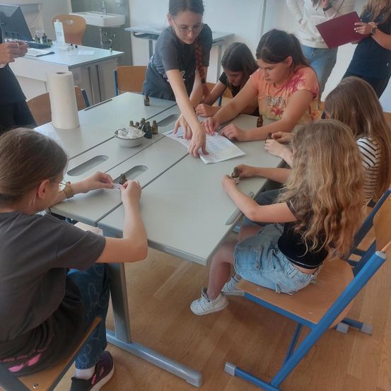 Eine Gruppe von Kindern sitzt um einen Tisch herum und arbeitet an einer Aktivität. Sie haben Flaschen, Schüsseln und Papiere vor sich. Ein Mädchen in der Mitte hält ein Papier und scheint etwas zu erklären. Ein anderes Mädchen schaut auf das Papier.