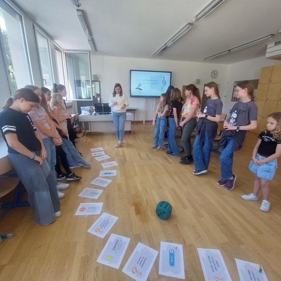 In einem Klassenzimmer bildet eine Gruppe junger Frauen einen Kreis um eine Frau in der Mitte, die ein Stück Papier in der Hand hält. An der Wand ist ein Beamer angebracht, der eine Präsentation zeigt. Fragebögen liegen auf dem Boden verstreut.