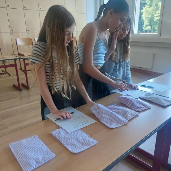 Drei Mädchen befinden sich in einem Klassenzimmer und arbeiten an einem Projekt. Sie sitzen an einem Tisch mit Papieren und Materialien. Ein Mädchen schreibt, während die anderen ein Stück Stoff inspizieren.