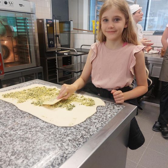 Ein kleines Mädchen in einem rosa Shirt macht Pizza in einer Küche. Sie hat einen Spatel und fügt dem Teig auf dem Tisch Toppings hinzu.