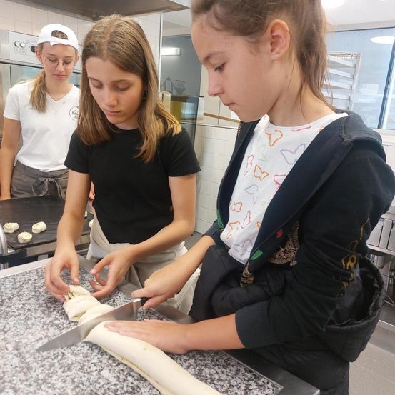 Drei Mädchen in einer Küche, eines schneidet Teig, während ein anderes zusieht. Ein drittes Mädchen steht hinter ihnen. Sie scheinen eine Backaufgabe zu erlernen oder zu üben.
