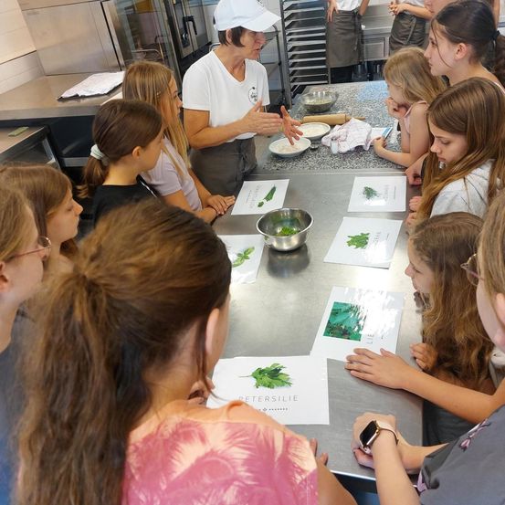 Eine Frau führt eine Gruppe von Mädchen in einer Küche an und zeigt ihnen, wie man Essen zubereitet. Auf dem Tisch liegen Papiere mit Rezepten und Zutaten.