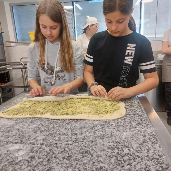 Zwei junge Mädchen bereiten Teig auf einer Marmorplatte vor. Eine trägt eine graue Kapuzenjacke, die andere ein schwarzes T-Shirt mit weißem Text. Eine Frau in einer weißen Haube steht hinter ihnen. Fenster und Regale sind im Hintergrund zu sehen.