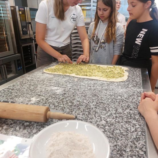 In einer Küche zeigt eine Frau zwei Mädchen, wie man Pizzateig vorbereitet. Der Teig liegt auf einer Arbeitsplatte mit einem Nudelholz und Mehl in einer Schüssel.