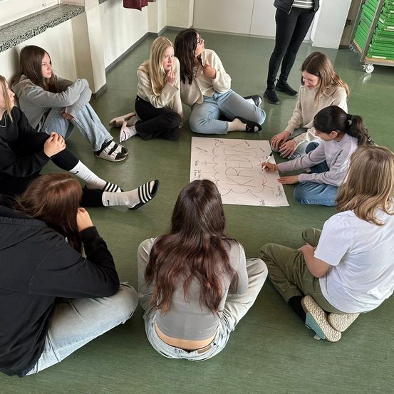 Mehrere Mädchen sitzen in einem Kreis auf dem Boden. Ein Mädchen zeichnet auf ein großes Blatt Papier. Einige tragen eine Brille. Ein Mädchen trägt einen schwarzen und weißen Schuh.