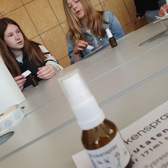 Zwei junge Mädchen arbeiten an einem Projekt am Tisch, wobei sie kleine Flaschen und Papier verwenden. Eine Person mit einem Stift ist in der Nähe.