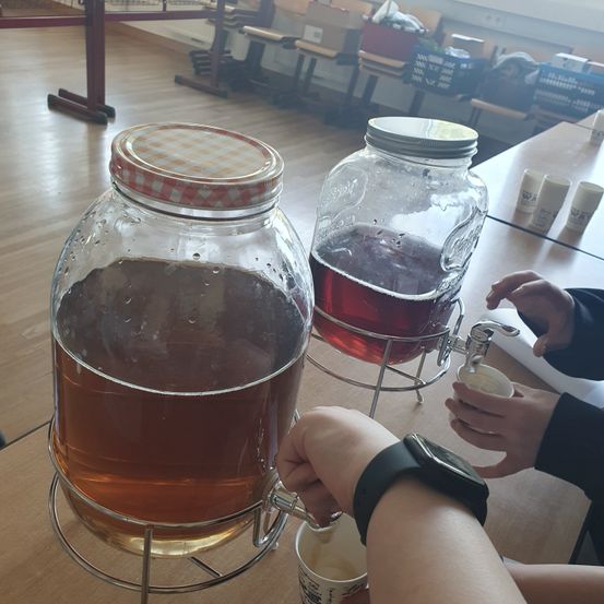 Zwei Glaskruken mit brauner Flüssigkeit stehen auf einem Tisch mit einer Tasse und einem Wasserhahn. Eine Person mit einer Uhr gießt aus der rechten Kruke.