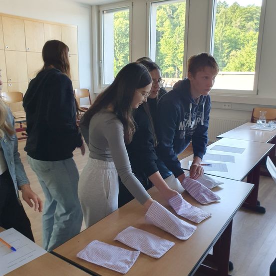 Schüler stehen in einem Klassenzimmer um einen Tisch herum und falten Stoffmasken. Zwei arbeiten am Tisch, während andere zusehen. Fenster mit Bäumen draußen sind dahinter.