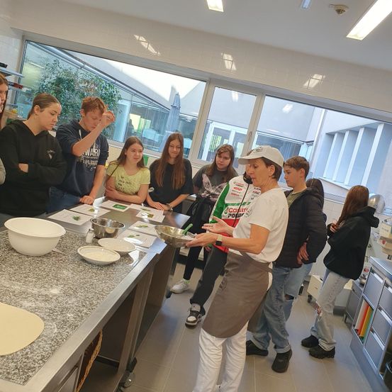 In einer Küche versammeln sich eine Gruppe von Schülern um eine Frau, die das Kochen demonstriert. Sie hält eine Schüssel und zeigt auf Zutaten auf dem Tresen. Sie beobachten aufmerksam.
