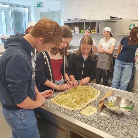 In einer Küche arbeiten mehrere Personen an einem Pizzateig auf einem Tresen. Eine Schüssel mit Toppings und ein Nudelholz sind in der Nähe.