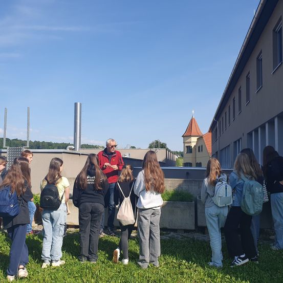 Eine Gruppe von Schülern versammelt sich auf einem Grasfeld und hört einem Mann in einer roten Jacke zu, wahrscheinlich ihrem Lehrer, der in der Nähe eines Gebäudes mit vielen Fenstern spricht.