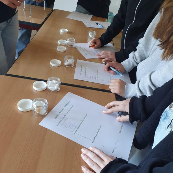 Eine Gruppe von Schülern in einem Klassenzimmer führt ein Experiment durch. Sie verwenden kleine Behälter mit Deckeln und schreiben auf Papier.