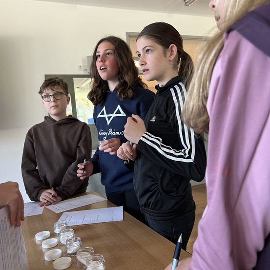 Drei junge Personen sind um einen Tisch versammelt und beschäftigen sich mit einer Aktivität. Eine Mädchen hält einen Stift und eine andere ein kleines Behältnis. Ein Junge schreibt auf ein Blatt Papier.