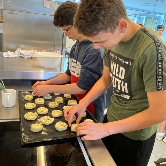 Zwei Jungen bereiten in einer Küche Gebäck zu. Einer trägt eine Brille und eine Kapuzenjacke, der andere ein grünes T-Shirt. Sie arbeiten an einem Tablett mit Gebäck, in der Nähe liegen eine Schüssel und ein Handtuch.