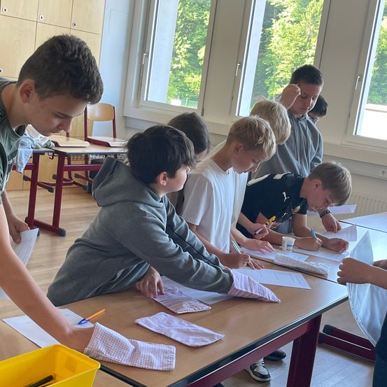 Eine Gruppe von Jungen sitzt um einen Tisch in einem Klassenzimmer und arbeitet mit Stiften an Papieren. Sie sind von Schreibtischen und Stühlen umgeben. Durch das Fenster sind Bäume zu sehen.