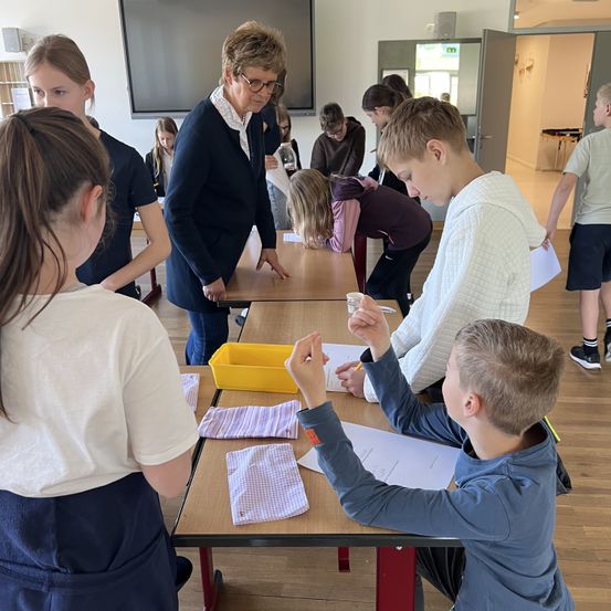 Mehrere Schüler sind um einen Tisch in einem Klassenzimmer versammelt. Eine Frau mit Brille und Blazer beobachtet sie. Papiere, ein gelbes Tablett und ein Becher stehen auf dem Tisch. Ein Junge hebt die Hand, während ein anderer auf Papier schreibt.