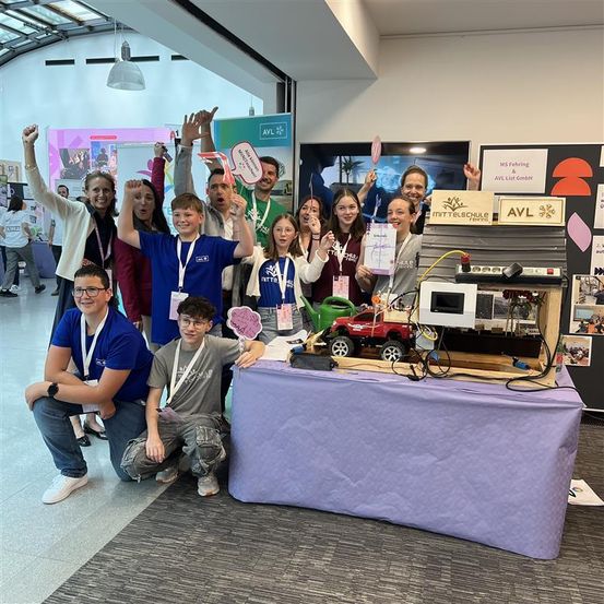 Eine Gruppe von Schülern und Lehrern posiert mit einem Modellauto und einer Spielzeuggießkanne auf einem Tisch. Sie alle haben Ausweiskarten und einige heben ihre Hände.