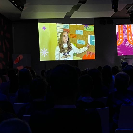 Eine junge Frau präsentiert auf einer Leinwand mit farbenfrohem Hintergrund, umgeben von Notizen und bunten Formen. Das Publikum sieht aufmerksam zu.
