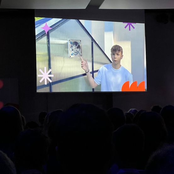 Ein Mann in einem weißen T-Shirt hält eine Präsentation vor einer Leinwand, auf der ein Gewächshaus mit einem Ventilator zu sehen ist.
