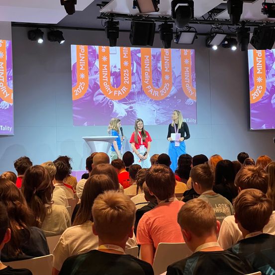Drei Frauen stehen auf der Bühne bei einer Veranstaltung, zwei mit Mikrofonen. Dahinter zeigt eine große Leinwand den Text 'MINTAIL 2025 FAIR'. Das Publikum sitzt vorne und hört aufmerksam zu.