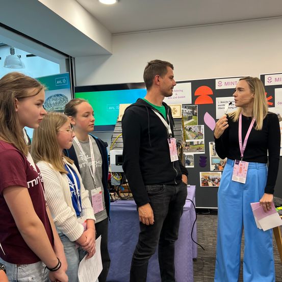 Ein Mann und eine Frau mit Mikrofon stehen vor einer Gruppe von Mädchen. Sie befinden sich in einem Raum mit einem Tisch und einem großen Bildschirm im Hintergrund.
