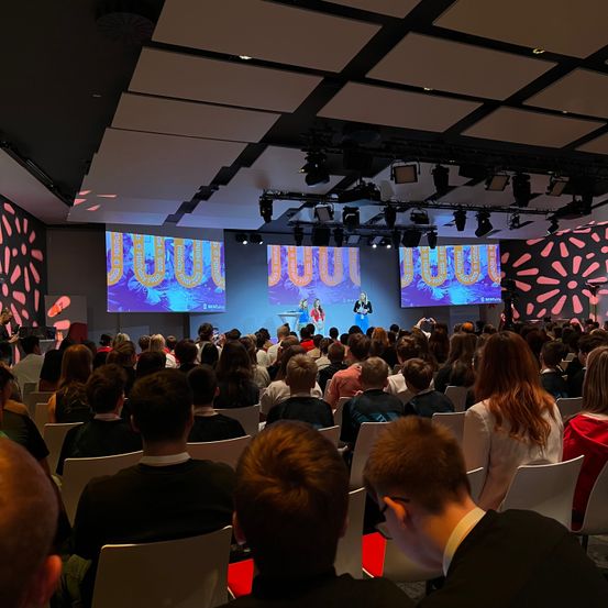 Ein großes Publikum, das in Reihen sitzt, beobachtet eine Präsentation auf einer Bühne mit zwei Personen und großen Bildschirmen dahinter. Der Raum hat eine moderne Einrichtung mit rosa Mustern an den Wänden.