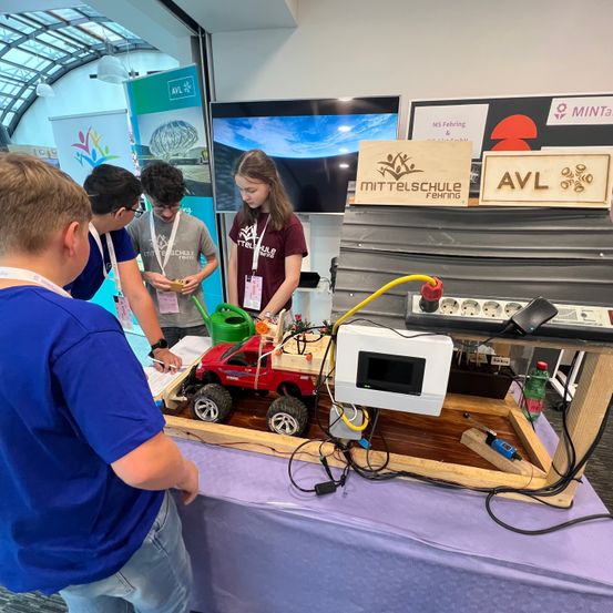 Vier Schüler untersuchen eine ferngesteuerte Autoanlage bei einer Schulveranstaltung. Das Auto steht auf einer Holzplattform mit Kabeln und Geräten. Ein Schüler schreibt auf ein Papier.
