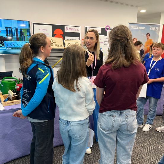 Eine Gruppe von Schülern und einer Frau stehen in einem Raum. Die Frau spricht zu den Schülern. Ein Tisch mit einigen Gegenständen steht vor ihnen. Dahinter ist ein Monitor an der Wand befestigt.