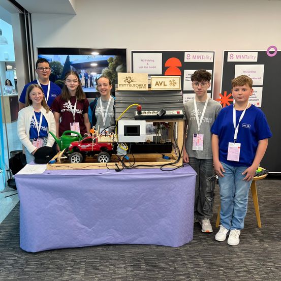 Eine Gruppe junger Menschen steht hinter einem Tisch mit einem kleinen Spielzeugauto und anderen Objekten zur Ausstellung. Sie tragen Namensschilder.