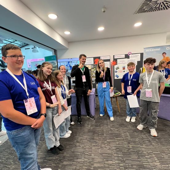 Eine Gruppe von Schülern und Erwachsenen steht in einem Raum mit Postern an der Wand. Die Schüler tragen Namensschilder und halten Papiere. Der Mann in der Mitte trägt eine schwarze Jacke und Turnschuhe.