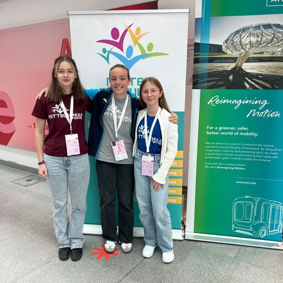 Drei junge Frauen stehen lächelnd für ein Foto vor einem Banner und posieren zusammen. Sie tragen Namensschilder mit Abzeichen. Dahinter befindet sich ein farbenfrohes Banner mit einem Baum und Text über die Neugestaltung der Bewegung.