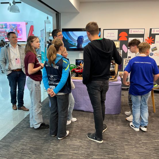 Eine Gruppe von Menschen ist in einem Raum versammelt und sieht sich eine Präsentation auf einem Monitor an. Ein Mann und eine Frau stehen vor einem Tisch.