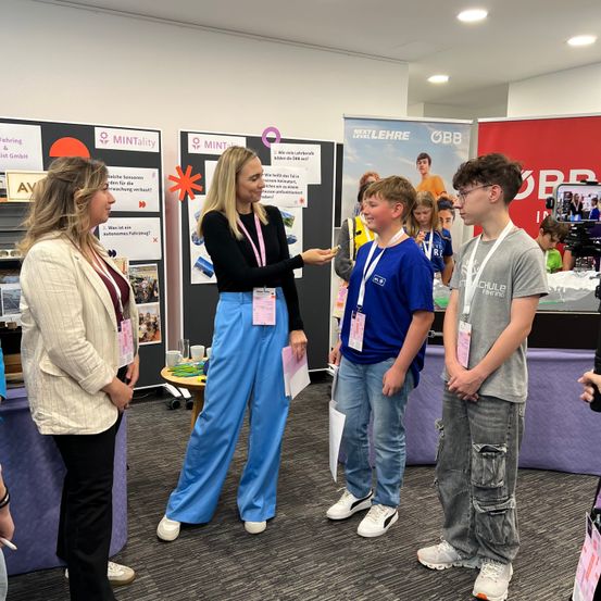 Eine Frau in einem weißen Blazer und schwarzen Hosen spricht mit einer Frau in einem schwarzen Shirt und blauer Hose. Sie werden von zwei Jungen begleitet, einer in einem blauen Shirt und der andere in einem grauen Shirt, bei einer Schulveranstaltung.